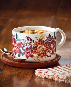 A white ceramic mug with a colorful floral pattern, filled with soup and placed on a wooden saucer with a spoon, set on a rustic wooden table.