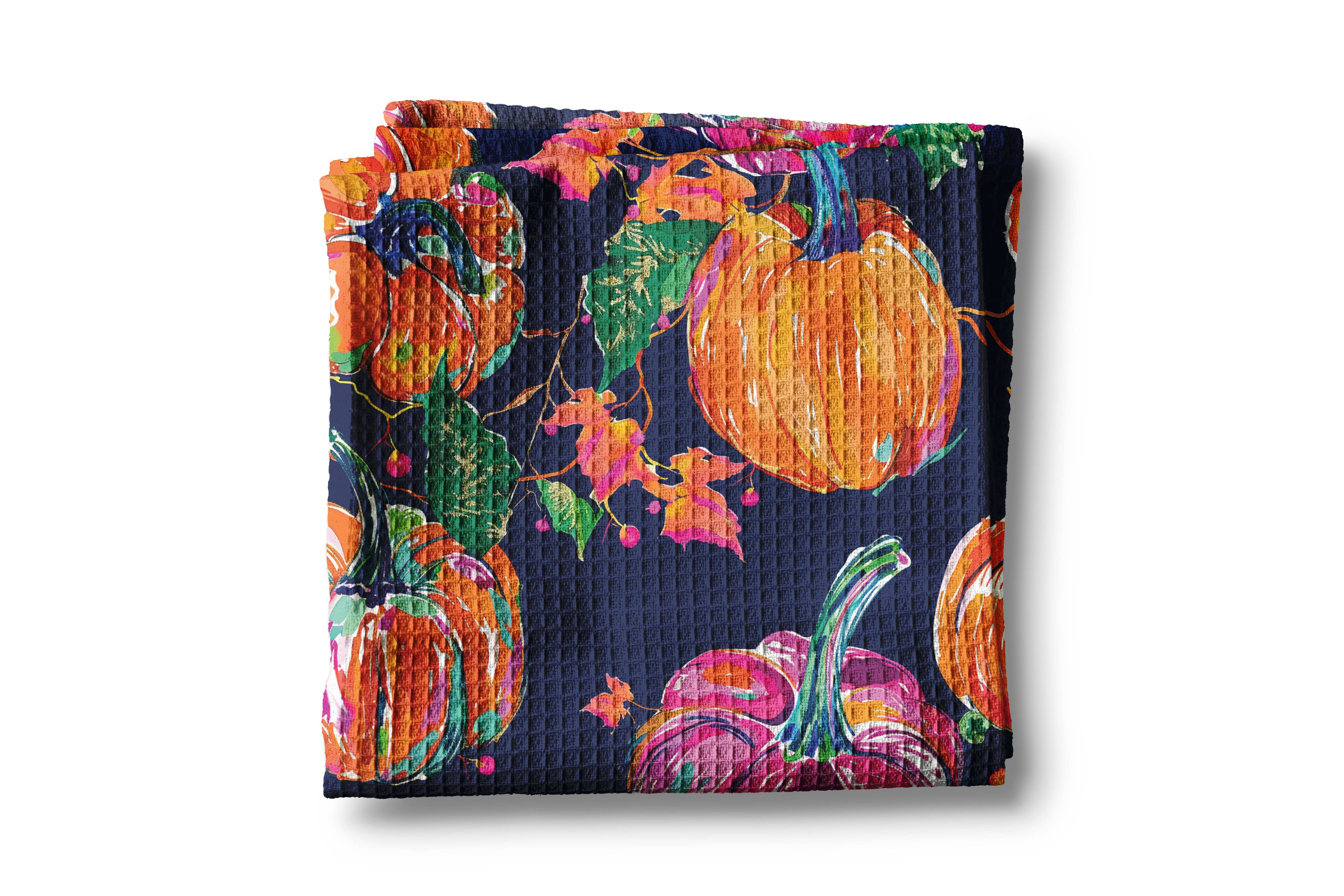A folded waffle-weave fabric item, likely a napkin or small towel, featuring a vibrant autumn pumpkin pattern on a dark navy blue background.